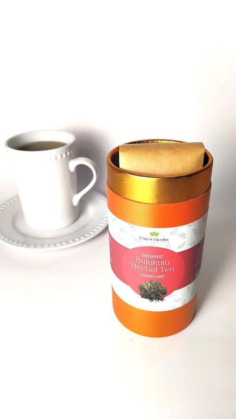 A white cup and saucer filled with tea sit beside an orange Forev'Herbs container labeled Organic Bulukutu Herbal Tea, with a rolled paper inside, highlighting a unique herbal tea experience against a plain white background.