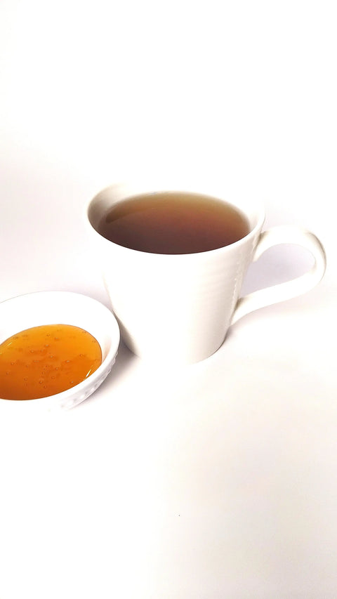 A white cup of Forev'Herbs Organic Bulukutu Herbal Tea sits beside a small white dish of honey, both arranged on a white surface with a minimalistic background.