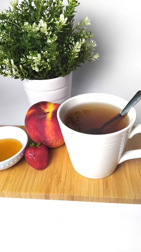A white cup of Forev'Herbs Organic Bulukutu Herbal Tea with a spoon sits on a wooden board, alongside a peach, strawberry, small dish of honey, and a green potted plant in the background.