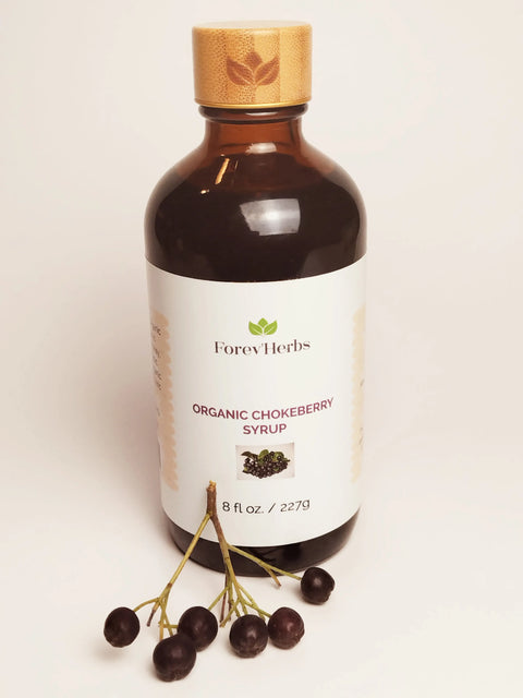 A brown bottle of Forev'Herbs Organic Chokeberry Syrup stands upright, featuring a green leaf logo and chokeberry image on the label, with antioxidant-rich aronia berries and stems arranged in front.