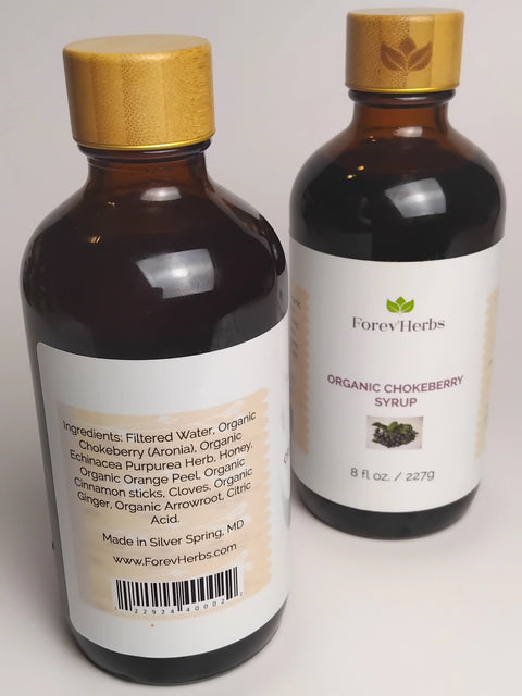 Two brown Forev'Herbs Organic Chokeberry Syrup bottles with wooden caps: one displays aronia berries, an antioxidant superfruit; the other features the ingredient list, website, and a small chokeberry image.