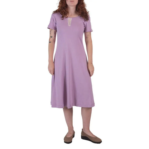 A woman with curly red hair models the knee-length, short-sleeve lavender Lake Shore Dress by Spiritex | Sustainable Apparel, featuring a lace detail at the neckline and paired with brown shoes against a white background.