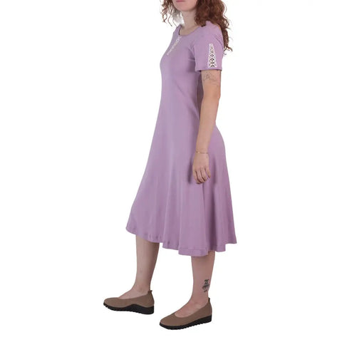 A woman stands in profile against a plain white background wearing the knee-length, short-sleeved Lake Shore Dress in lavender organic cotton by Spiritex | Sustainable Apparel, paired with brown flats.
