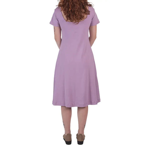 A woman with curly red hair faces away, wearing a short-sleeved, knee-length lavender Lake Shore Dress by Spiritex | Sustainable Apparel, made from organic cotton. She pairs it with brown shoes against a white background.