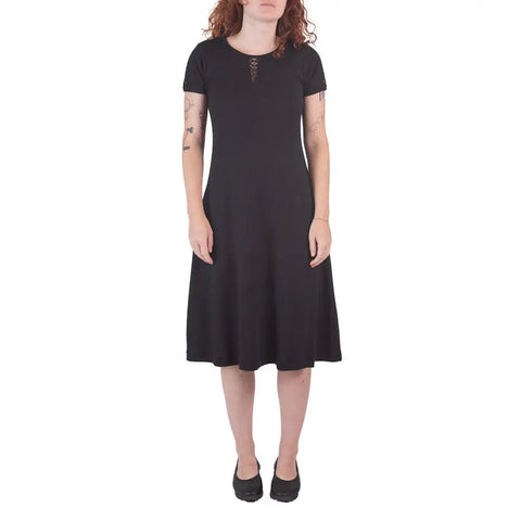 A woman with curly red hair wears the Spiritex | Sustainable Apparel Lake Shore Dress, featuring short sleeves and a lace-up neckline, paired with black shoes, standing against a white background.