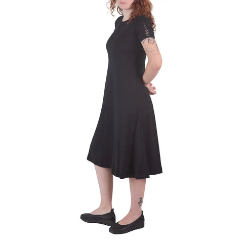 A woman with curly hair models the knee-length Lake Shore Dress by Spiritex | Sustainable Apparel, standing in a side pose with her arms behind her back, showing arm and ankle tattoos, against a white background.