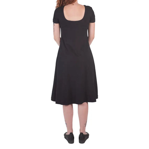 A woman with curly hair and arm tattoos stands facing away in a black Spiritex | Sustainable Apparel Lake Shore Dress featuring short sleeves and a scoop back, paired with black shoes against a plain white background.