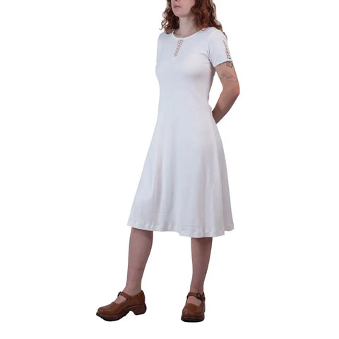 A woman with wavy hair stands against a white background, wearing Spiritex | Sustainable Apparels knee-length, short-sleeve Lake Shore Dress made from organic cotton and brown shoes, with her arms behind her back.