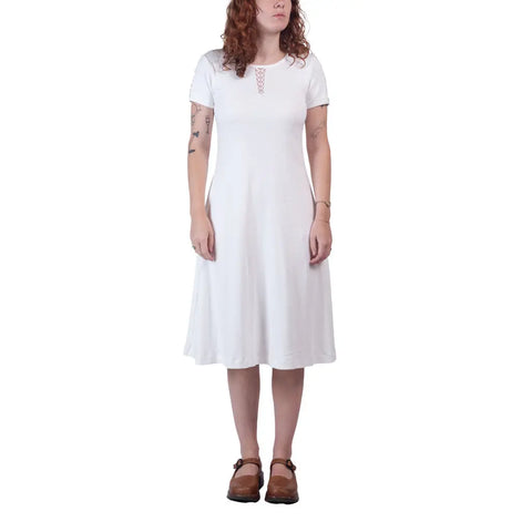 A woman with curly reddish hair wears a knee-length, short-sleeved white Lake Shore Dress by Spiritex | Sustainable Apparel and brown clogs, standing against a white background.
