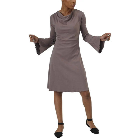A woman wears the Spiritex Grove Dress—an organic cotton, taupe A-line with long sleeves and wide flared cuffs. She poses with arms raised and legs apart in black flats against a white background.