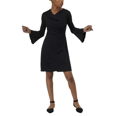 A person models the Spiritex | Sustainable Apparel Grove Dress—a knee-length black A-line piece with long flared sleeves—against a white background, styled with black ankle-strap shoes and their head turned to the side.