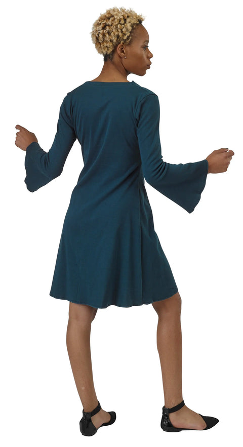 A woman with short curly hair models the Grove Dress by Spiritex | Sustainable Apparel—an organic cotton, long-sleeved teal A-line dress—paired with black ankle-strap flats. She stands facing away, turning her head left.