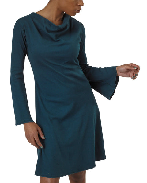 A woman models the Grove Dress by Spiritex | Sustainable Apparel, featuring long sleeves, a loose cowl neckline, slightly flared sleeves, and crafted from teal organic cotton. She poses with one arm bent, gazing to the side.