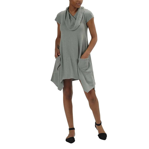 A woman wears the Colette Drape Dress by Spiritex | Sustainable Apparel—a loose-fitting gray dress with short sleeves, cowl neckline, asymmetrical hem, and two front pockets—paired with black ankle-strap flats.