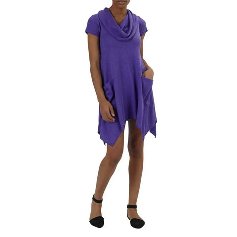 A woman wears the Colette Drape Dress by Spiritex | Sustainable Apparel, a loose-fitting purple bamboo jersey dress with a cowl neck and pockets, paired with black ankle-strap flats and one hand in her pocket.