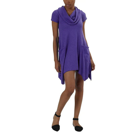 A person models the short-sleeved, purple Colette Drape Dress by Spiritex | Sustainable Apparel, featuring a cowl neck, asymmetrical hem, and large front pockets, paired with black ankle-strap flats against a white background.