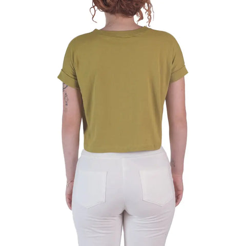 A woman with curly hair tied up and visible arm tattoos faces away, wearing Spiritex | Sustainable Apparel’s Roan Crop Top in olive green, made from 100% organic cotton, paired with high-waisted white pants.