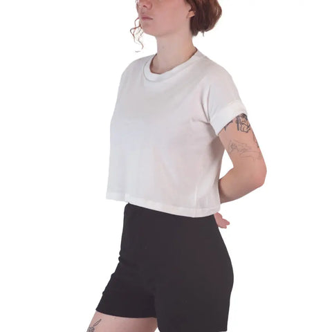 A person with short curly hair and tattoos models the Roan Crop Top by Spiritex | Sustainable Apparel, styled with black high-waisted shorts, standing hands behind their back against a white background—showcasing sustainable fashion.