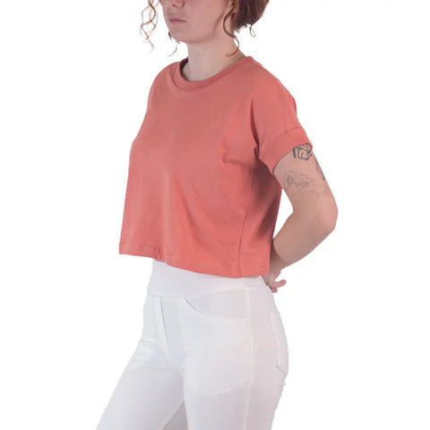 A person with curly hair wears the coral Roan Crop Top by Spiritex | Sustainable Apparel, made from 100% organic cotton, paired with white high-waisted pants. They stand with hands behind their back, revealing an upper arm tattoo against a white background.