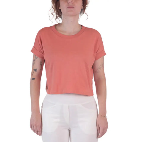 A woman in a short-sleeved, coral Roan Crop Top by Spiritex | Sustainable Apparel, made from 100% organic cotton, and high-waisted white pants stands facing forward against a white background, her tattooed forearms relaxed at her sides.