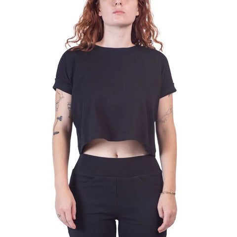 A person with long curly hair is wearing a black Roan Crop Top by Spiritex | Sustainable Apparel and black pants. Their tattooed arms are relaxed by their sides, against a plain white background to highlight their sustainable style.