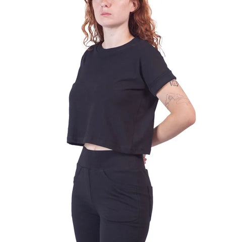 A person with wavy red hair wears the Roan Crop Top by Spiritex | Sustainable Apparel in black organic cotton, paired with black pants, posing against a white background with one arm behind their back and a forearm tattoo visible.