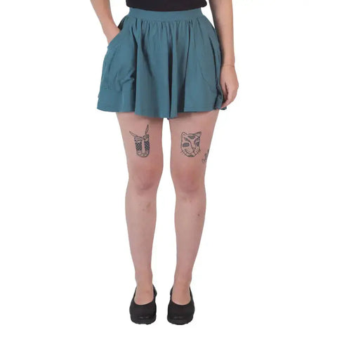 A person wearing Spiritex Drape Shorts in teal, paired with a black top and black shoes, stands with hands in pockets. Tattoos—a moth on the left thigh and an owl on the right—are visible. Photo is from shoulders down.