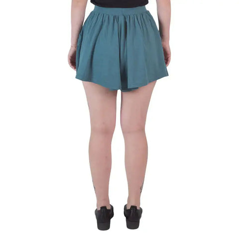A person stands with their back to the camera, wearing Spiritex | Sustainable Apparel Drape Shorts in teal, a black top, and black shoes. Their tattooed ankles stand out against a plain white background—a relaxed summer look.