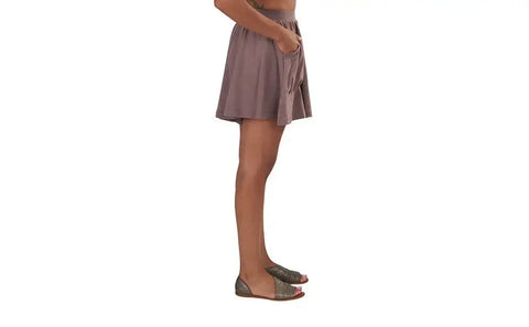 A person stands sideways in Spiritex | Sustainable Apparel Drape Shorts and green open-toed flats, hands in pockets, against a white background—an effortless choice for a stylish summer wardrobe.