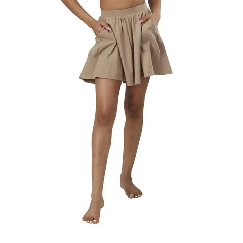 A person stands barefoot with hands in the pockets of tan, flared, knee-length Drape Shorts by Spiritex | Sustainable Apparel against a white background. The upper body and face are not visible.
