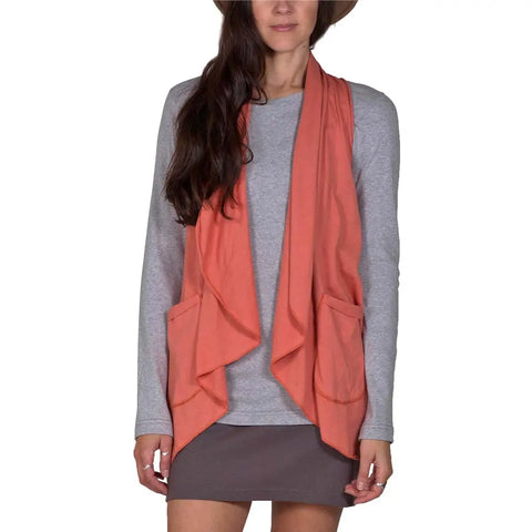 A woman stands against a white background wearing a gray long-sleeve shirt, gray skirt, and the Spiritex Ultra Lightweight Draped Vest in coral with front pockets. Her face is partly hidden by her hair and hat.