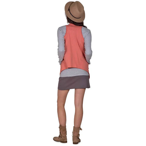A person stands facing away, wearing the Spiritex Ultra Lightweight Draped Vest in coral organic cotton over a gray long-sleeve shirt, paired with a short gray skirt and brown hat and boots, set against a plain white background.