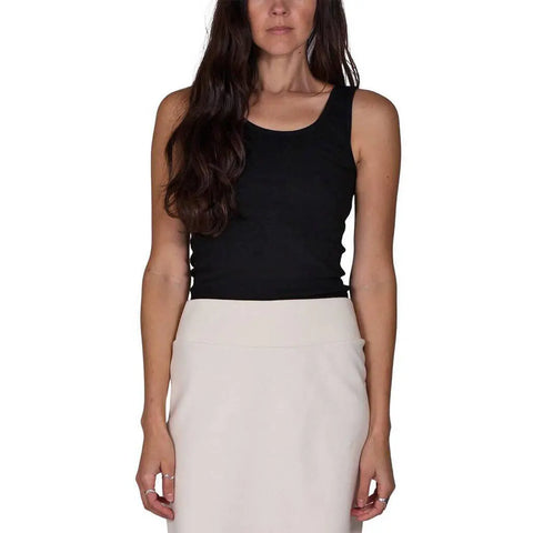 A woman with long dark hair wears the Spiritex Classic Tank, a sleeveless black top, paired with a high-waisted, light-colored 100% organic cotton skirt, standing against a plain white background with her arms relaxed by her sides.