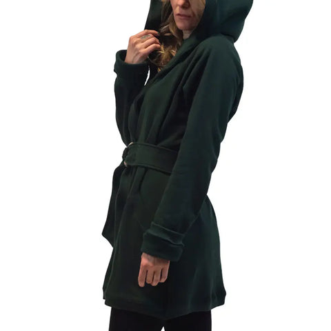 A woman stands sideways against a white background, holding the hood of the Spiritex Fleece Wrap Jacket—an organic cotton, dark green belted coat—from Spiritex | Sustainable Apparel.