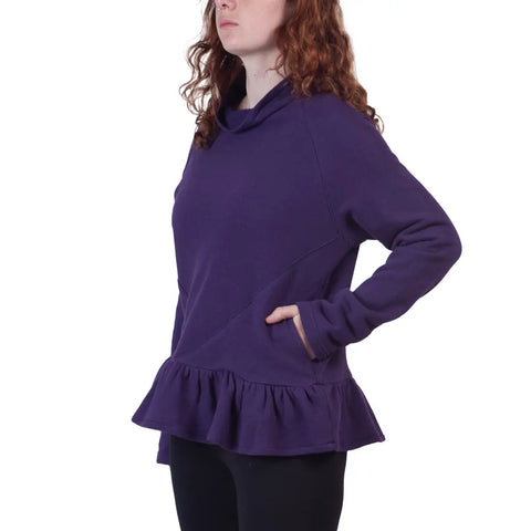 A woman with long curly red hair stands in profile against a white background, wearing the Charlotte Street Sweatshirt by Spiritex | Sustainable Apparel—an oversized turtleneck with a ruffled hem and one hand in the side pocket.