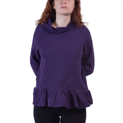 A person with wavy, shoulder-length hair wears the Spiritex | Sustainable Apparel Charlotte Street Sweatshirt in purple with long sleeves, a wide collar, and ruffled hem, paired with black pants. Their arms are behind their back; background is plain white.