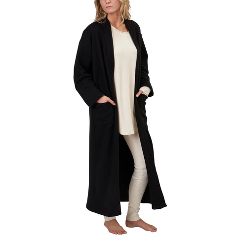 A barefoot woman in cream pajamas and a Spiritex | Sustainable Apparel Full-Length Fleece Robe stands with her hands in the robe’s pockets against a white background.