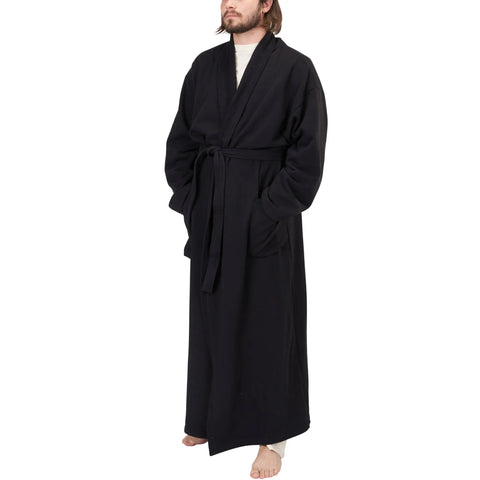 A person stands barefoot in the Spiritex Full-Length Fleece Robe, a sustainable black organic cotton garment with pockets and a waist belt, against a plain white background.
