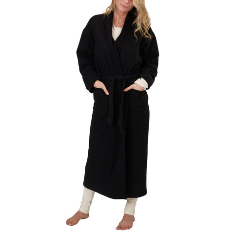 A woman stands barefoot in a Spiritex Full-Length Fleece Robe—black, organic cotton, with a belted waist and pockets—worn over white pants and a white top. She has wavy blonde hair. Brand: Spiritex | Sustainable Apparel.