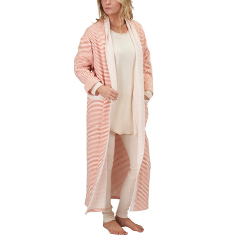 A woman stands barefoot, slightly to the side, holding open a Spiritex | Sustainable Apparel Full-Length Fleece Robe in light pink and white stripes, worn over cream pajamas.