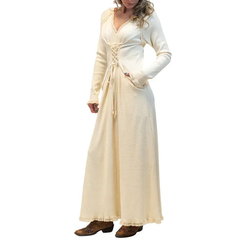 A woman stands against a plain white background wearing the Spiritex | Sustainable Apparel Gwynne Dress, a long cream-colored, corset-inspired piece with lace-up details and long sleeves, paired with brown shoes and one hand in her pocket.