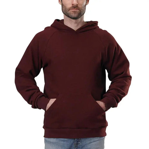 A man stands against a white background wearing the Spiritex | Sustainable Apparel Heavyweight Fleece Hoodie in maroon with a lined hood and front pocket, paired with light blue jeans.