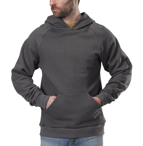 A man stands against a white background, wearing a Spiritex | Sustainable Apparel Heavyweight Fleece Hoodie in gray with a lined hood and front pocket, layered over an organic cotton mustard shirt and paired with blue jeans.