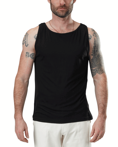 A man models the Spiritex | Sustainable Apparel Jersey Tank in black, paired with white pants, revealing his tattooed arms. He stands facing forward against a plain white background, with only the lower part of his face visible.