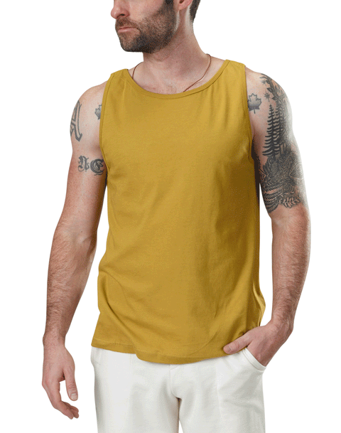A bearded, tattooed man wears a sleeveless mustard yellow Spiritex Jersey Tank made from 100% organic cotton and white pants, standing against a plain white background with one hand in his pocket, looking to his left.