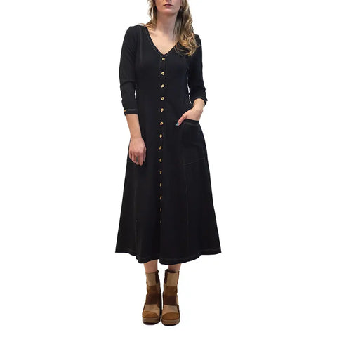 A woman wears the Spiritex Leaf Dress, a black, long-sleeve, button-down autumn dress with front pockets, paired with brown platform shoes. She stands against a white background with loose, wavy hair and her face partially out of frame.