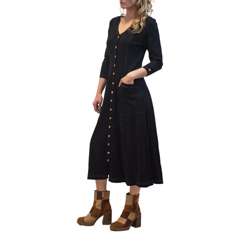 A woman wears the Spiritex Leaf Dress, a long-sleeve, button-down black autumn dress with pockets, paired with brown heeled ankle boots, standing against a white background.