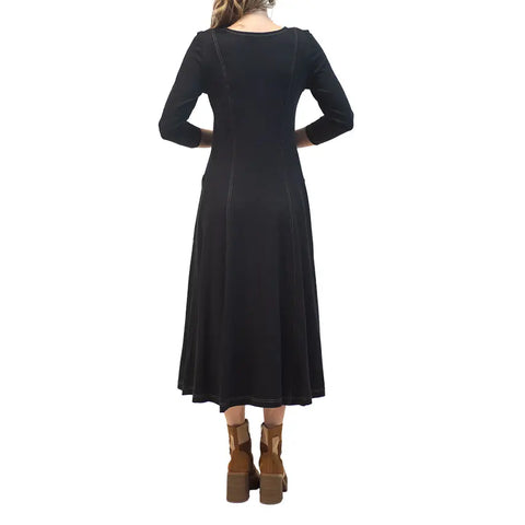A woman faces away in the Spiritex Leaf Dress, an ankle-length black organic cotton piece with long sleeves and subtle seam lines, paired with brown heeled ankle boots. The background is white.