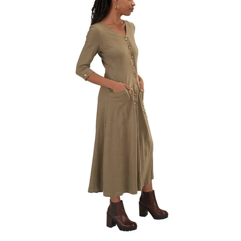 A woman wearing the Spiritex Leaf Dress—an organic cotton, long khaki dress with wooden leaf buttons, three-quarter sleeves, and front pockets—paired with dark brown platform boots, stands against a white background.