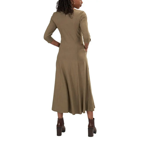 A woman with curly hair faces away, modeling the Spiritex Leaf Dress—a calf-length, olive green organic cotton dress with wooden leaf buttons and three-quarter sleeves—paired with brown heeled boots against a white backdrop.